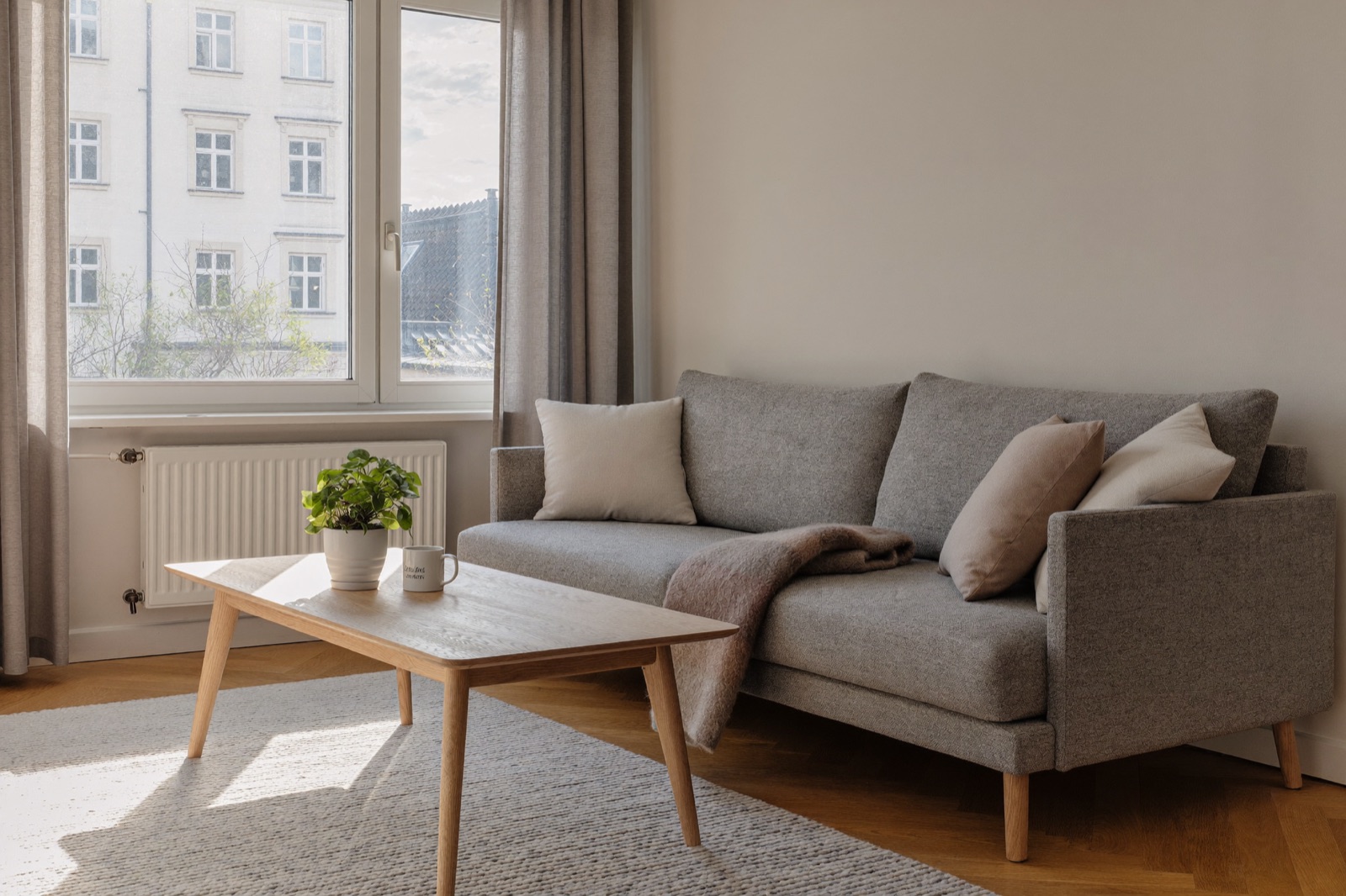 Wygodna szara sofa z poduszkami w jasnym salonie, obok drewniany stolik kawowy i duże okno